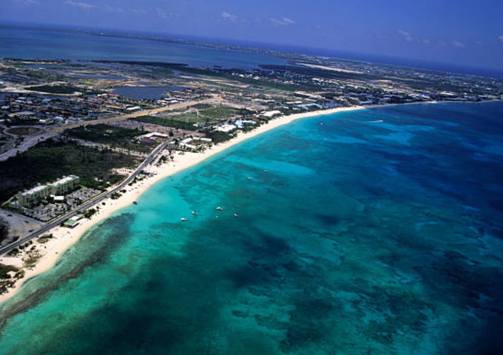 Aerial view of Seven Mile Beach Grand Cayman with white sand, turquoise reef water, resorts along the coast and excursion boats near shore