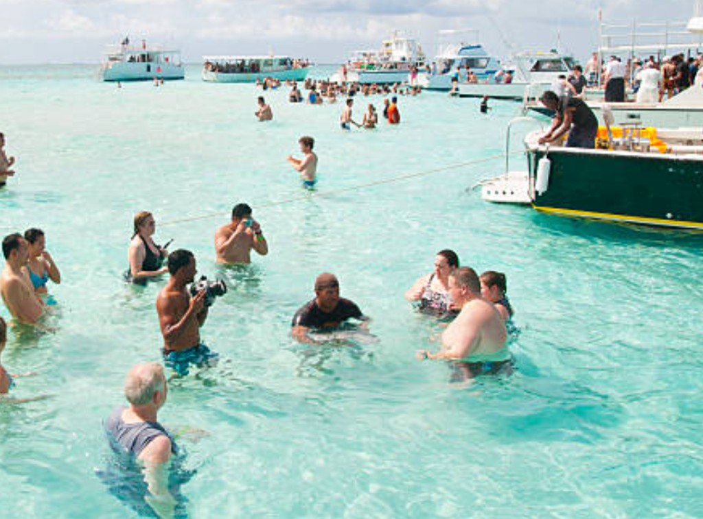 Tourists with excursion guides interacting with stingrays in shallow turquoise water at Stingray City sandbar Grand Cayman with tour boats anchored nearby